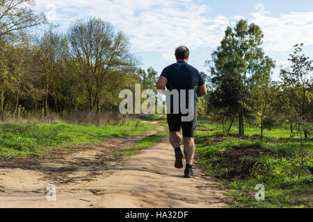 Big belly man jogging , exercising, doing cardio in the park , slightly overweight, loosing weight. On a lawn of green grass between trees without lea Stock Photo