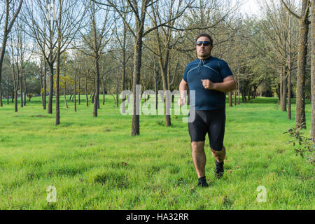 Big belly man jogging , exercising, doing cardio in the park , slightly overweight, loosing weight. On a lawn of green grass between trees without lea Stock Photo