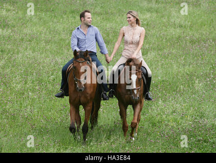 Horseman and horsewoman on horseback riding hand in hand through a green meadow Stock Photo
