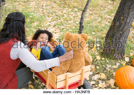 Mother playing with daughter and teddy bear in wagon in autumn leaves