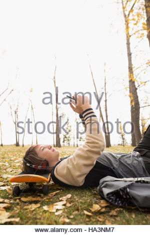 Boy with headphones and cell phone laying on skateboard in autumn park