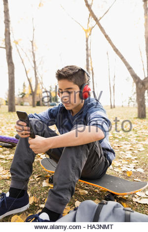 Tween boy with headphones and cell phone sitting on skateboard in autumn park