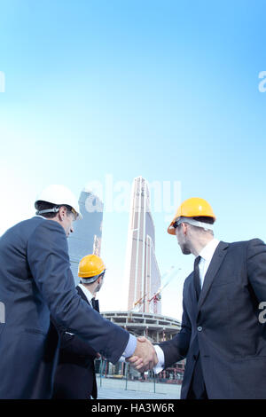Architects wearing helmets shaking hands at skyscrapers background Stock Photo