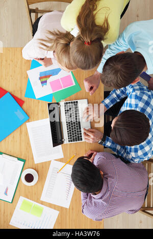 Businesspeople sitting at table looking at laptop screen, discussing financial documents with diagrams, top view Stock Photo