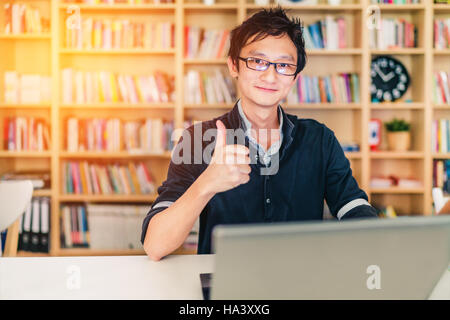 Young adult Asian man with laptop, thumbs up ok sign, home office or library scene, bookshelf with clock blur background with copy space, success or t Stock Photo