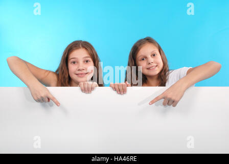 Two happy smiling beautiful young girl showing blank signboard or copyspace for slogan or text, isolated on blue background Stock Photo