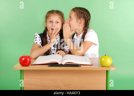 Two school girl sharing secrets  sitting at a desk from book isolated on green Stock Photo