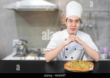 Asian young man chief in white uniform offers to you delicious pizza. Man chief preparing pizza in kitchen. Stock Photo