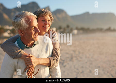 Portrait of happy mature man carrying his beautiful wife on his back at the beach. Senior couple enjoying their vacation at the sea shore. Stock Photo