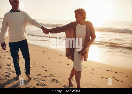 Outdoor shot of romantic senior couple walking along the sea shore holding hands. Senior man and woman walking on the beach together at sunset. Stock Photo