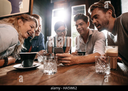Young friends looking at smart phone while sitting in cafe. Mixed race people sitting at a table in restaurant using mobile phone. Stock Photo