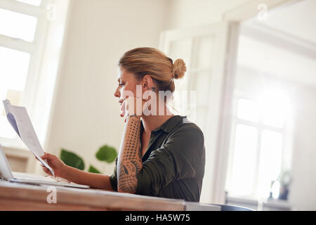 Side portrait of young female sitting at table reading documents. Woman going through contract papers at home office. Stock Photo