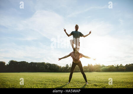 Strong young couple doing acrobatic yoga exercise in park. Man standing on back of woman and balancing. Stock Photo