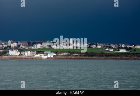 Deep blue stormy sky over Milford Haven with sunlight on church, houses and a patch of bright green grass over a cooler sea Stock Photo