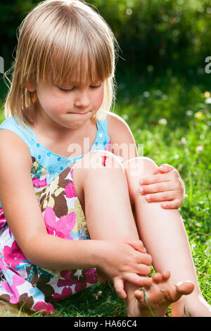 Little girl sitting on a lawn in a summer garden looking at injury on her knee with pursed lips holding back the tears Stock Photo