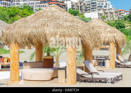 Wooden canopy made of dried palm leaves over sandy beach sunbeds in public area of hotel, nobody. Stock Photo