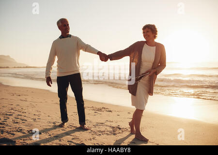 Full length outdoor shot of loving mature couple walking along the beach holding hands. Senior man and woman walking on the sea shore at sunset. Stock Photo