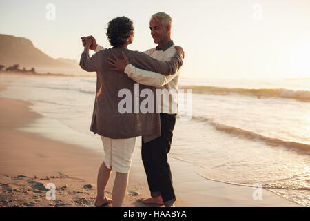 Senior man and woman couple dancing at sunset on the sea shore. Mature couple enjoying a romantic day at the beach. Stock Photo