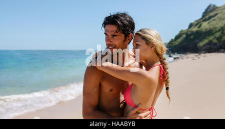 Affectionate young couple embracing on the beach and looking at the sea. Romantic young couple together on sea shore looking away. Stock Photo