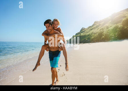 Portrait of man carrying girlfriend on his back along the sea shore. Man giving piggyback ride to girlfriend on the beach. Stock Photo
