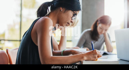 Close up shot of young woman writing notes with classmates studying in background. Students learning in college library. Stock Photo