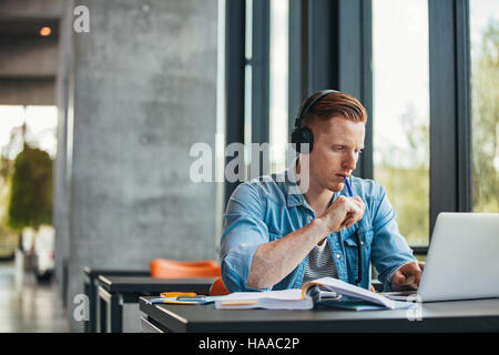 Male student sitting at table looking at laptop and thinking. Young man surfing internet on laptop at university library. Stock Photo