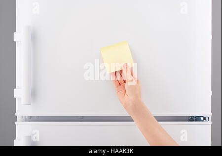 Female hand holding blank light yellow sticky paper note on white refrigerator door Stock Photo