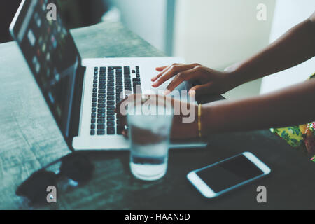 Side view of woman hands using laptop on a stone table with glass of water, smartphone and sunglasses on it, dark cafe interior Stock Photo