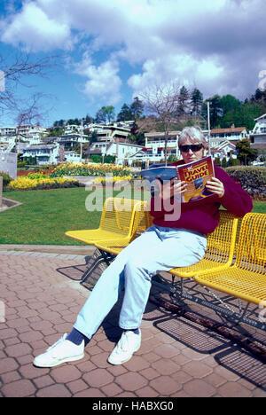 Middle-Aged Woman sitting on a Bench Outside and reading a Book, White Rock, BC, British Columbia, Canada Stock Photo