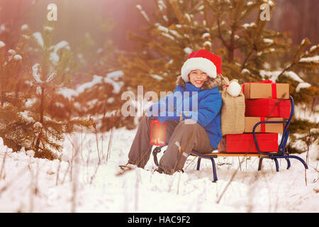 Cute little boy in Santa hat holding Christmas lantern and carries a wooden sled full of gift boxes in snowy forest. Xmas, snow and winter fun for fam Stock Photo