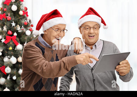 Happy seniors wearing santa hats looking at a tablet in front of a christmas tree isolated on white background Stock Photo