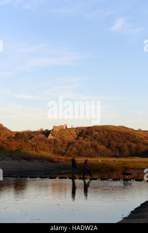 a couple with their dog cross the stepping stones at  Three cliffs bay, Gower, Wales at sunset . Stock Photo