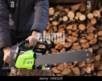 CLoseup of hands of a man holding an Electric Cordless chainsaw in front of stacked firewood in the background Stock Photo