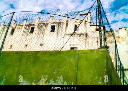 Green wall of sports ground topped with wire fence  in central Havana dilapidated building behind against blue sky white clouds Stock Photo