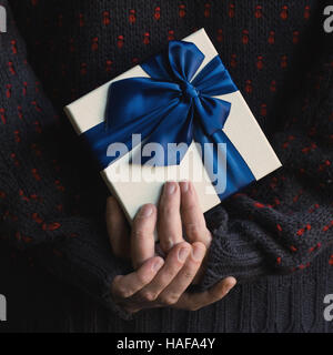 Close up of a man in knitted dark sweater holding a white present gift box with blue ribbon behind his back Stock Photo