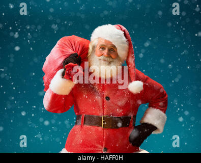 Smiling Santa Claus carrying big bag full of gifts to children on blue background. Merry Christmas & New Year's Eve concept. Stock Photo