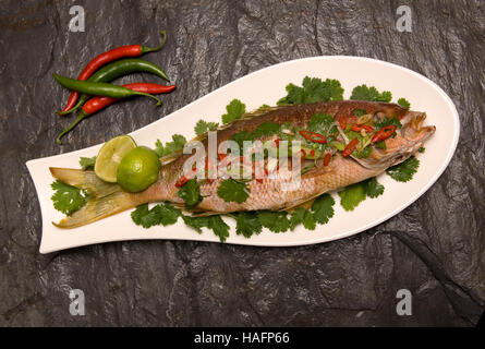 A whole cooked seabass on a fish-shaped white dish sitting on a dark slate background. Stock Photo