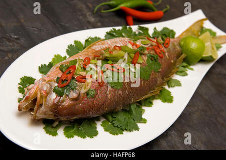 A whole cooked seabass on a fish-shaped white dish sitting on a dark slate background. Stock Photo