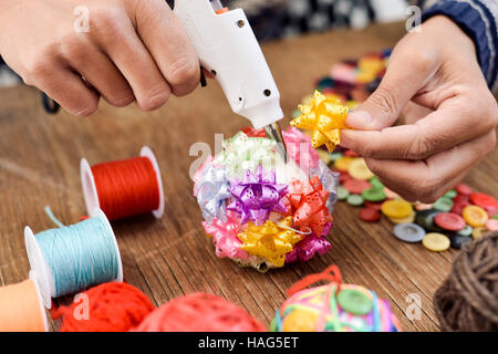 closeup of a young man making a handmade christmas ball with ribbon bows of different colors, using a hot glue gun, and a pile of different haberdashe Stock Photo