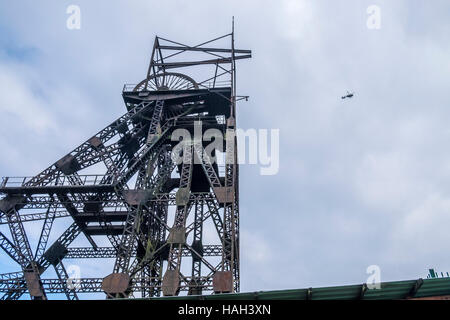 Pit head winding gear for lowering the miners cage into the mine, with a helicopter flying overhead. Stock Photo