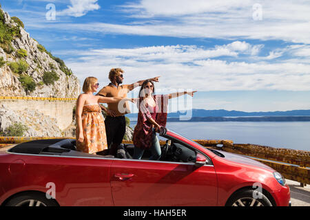 Group of happy young people waving and pointing fingers from the red convertible. Stock Photo