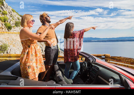 Group of happy young people standing in red convertible and pointing hands. Stock Photo