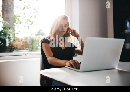 Thoughtful young woman sitting in kitchen and working on laptop. Female surfing internet on laptop at home. Stock Photo