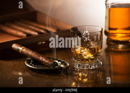 Glass of whiskey with ice cubes and smoking cigar on wooden table Stock Photo