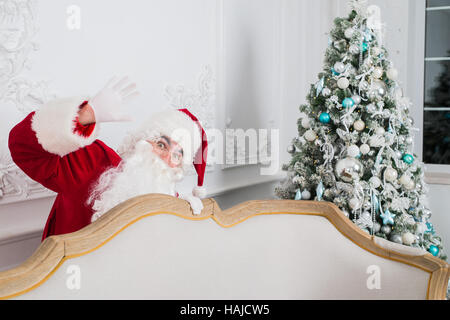 Santa Claus standing behind a Christmas tree and sofa looking at the camera Stock Photo