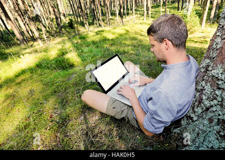 man is sitting against a tree in the forest, working with his laptop Stock Photo