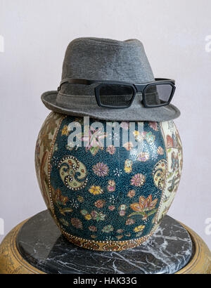 Gray trilby hat wearing sunglasses over antique decorated Chinese ceramic jar vase over antique black marble table Stock Photo