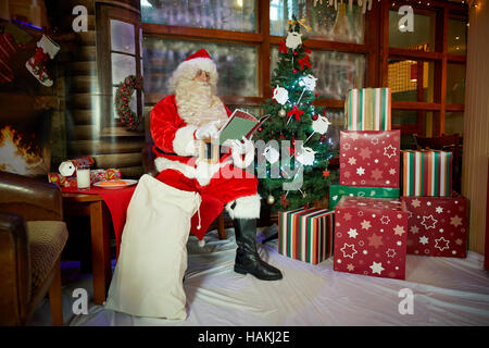 Santa in grotto christmas display   lighted christmas tree, presents,fireplace,stockings gifted wrapped reading costume good sack red in pub Santa Cla Stock Photo