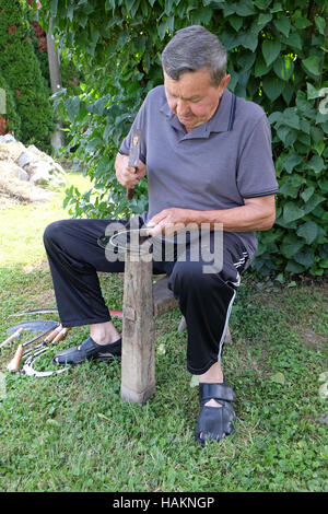 Farmer with hammer and iron tool on the tree stump is sharpening his scythe in Nedelisce, Croatia Stock Photo