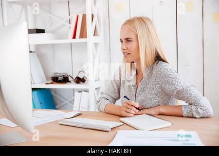 Portrait of a casual young businesswoman making notes while looking at computer monitor Stock Photo